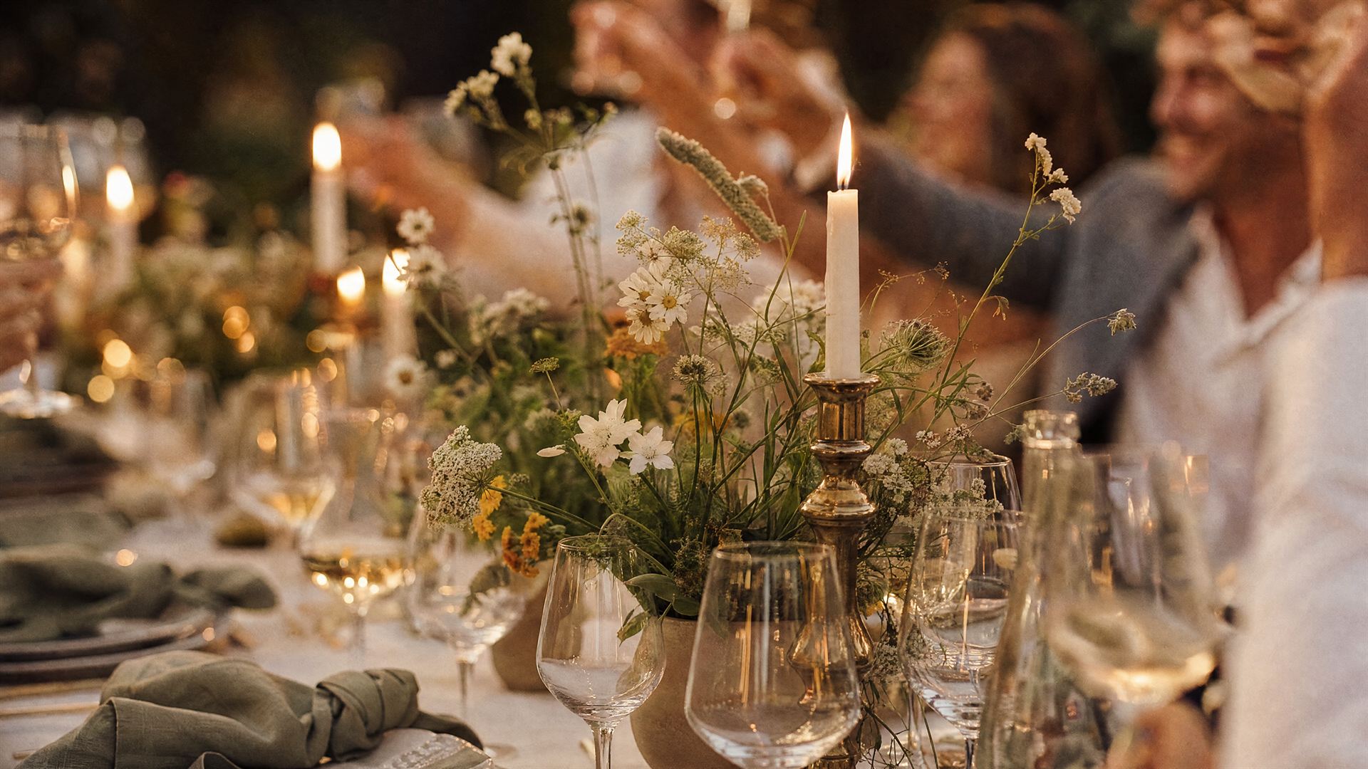 Dukat långbord med levande ljus och blomsterarrangemang medan gäster höjer sina glas i kvällsljuset – inspirationsbild